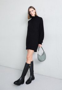 Robe-pull noire en maille XXL, longueur genoux, associée à des bottes en caoutchouc noires montant jusqu'aux genoux et un sac à main vert texturé. Design minimaliste.