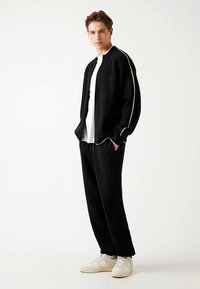 Giubbino bomber in maglia nera con una striscia bianca, indossato sopra una camicia bianca, abbinato a pantaloni neri e sneakers bianche, in piedi contro uno sfondo chiaro.