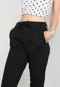 Pantaloni neri a vita alta con vita regolabile, dotati di tasche laterali e una texture liscia. Accentuati da un bracciale d'argento al polso.