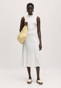 Witte mouwloze jurk met een aansluitende bovenkant en een uitlopende rok, gedragen met crèmekleurige sandalen. Het model draagt een zachte, gele schoudertas.