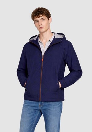 Giovane uomo che indossa una giacca con cappuccio blu navy con zip frontale e tasche, abbinata a jeans azzurri, in piedi davanti a uno sfondo semplice.