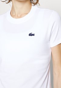 T-shirt en coton blanc à manches courtes, présentant un logo Lacoste bleu marine sur la poitrine gauche. Texture lisse et encolure ronde classique.