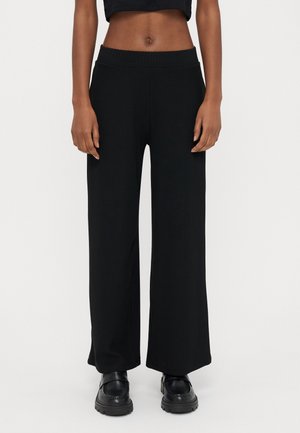 Vrouw die een zwarte, loszittende geribde broek met hoge taille en zwarte chunky loafers draagt, weergegeven van taille tot voeten tegen een effen achtergrond.