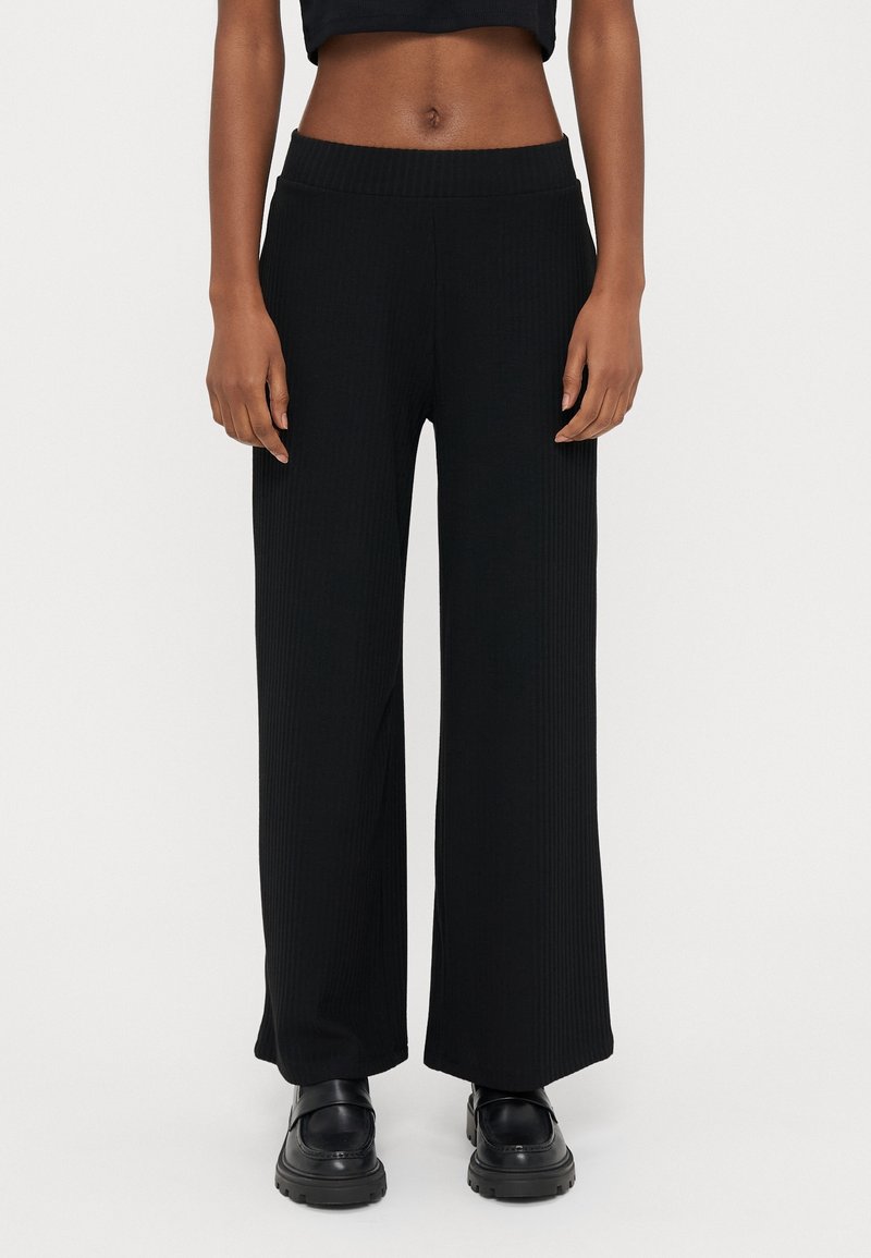 Femme portant un pantalon noir côtelé, ample et taille haute, ainsi que des mocassins noirs épais, montrée de la taille aux pieds sur un fond uni.