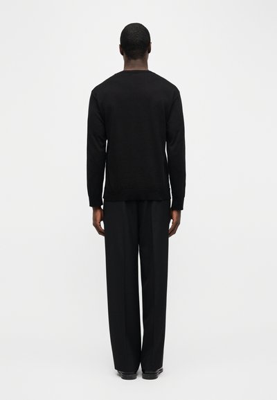 Άνδρας που στέκεται με την πλάτη γυρισμένη, φορώντας μαύρο πουκάμισο με μακριά μανίκια, μαύρο παντελόνι φαρδύ και μαύρα παπούτσια, μπροστά σε απλό λευκό φόντο.