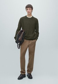 Maglione verde scuro texturizzato, colletto beige, pantaloni marroni e scarpe nere. Modello che tiene una giacca di pelle marrone sulla braccia.