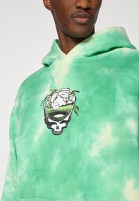 Πράσινη hoodie tie-dye από βαμβάκι, με γραφικό κρανίο με πράσινα στοιχεία και κυκλικό σχέδιο στο στήθος.