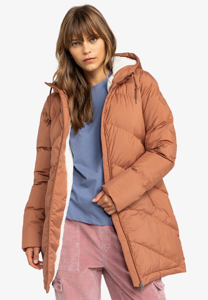 Bruine pufferjack met kap, voorzien van een pluche witte voering en diagonale gestikte afwerking. Gecombineerd met een blauwe shirt en roze broeken.