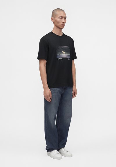Un homme se tient debout, portant un t-shirt noir à motif graphique, un jean bleu ample et des baskets blanches sur un fond blanc uni.