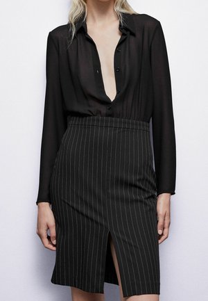 Zwarte transparante blouse met een diepe V-hals en knoopsluiting aan de voorkant, gecombineerd met een zwarte rok met fijne strepen en een zijsplit.