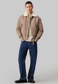 Chaqueta bomber marrón con un cuello de borreguito crema, suéter beige y vaqueros baggy azul oscuro. Zapatos negros y detalle de cremallera en la chaqueta.
