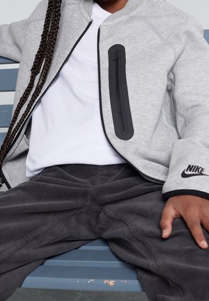 Enfant portant une veste Nike grise avec une poche zippée noire et un logo, un t-shirt blanc et un pantalon en velours côtelé foncé assis sur un banc bleu.