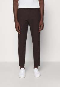 Pantalones oscuros de corte entallado en marrón, con frente plano, cierre de botón y dobladillos enrollados. Combinados con zapatillas blancas.