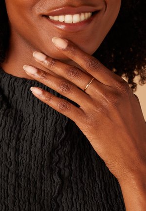 Bague en or sur un doigt reposant sur un tissu noir texturé. Teint de peau lisse, ongles naturels avec un vernis léger et un sourire chaleureux visible.