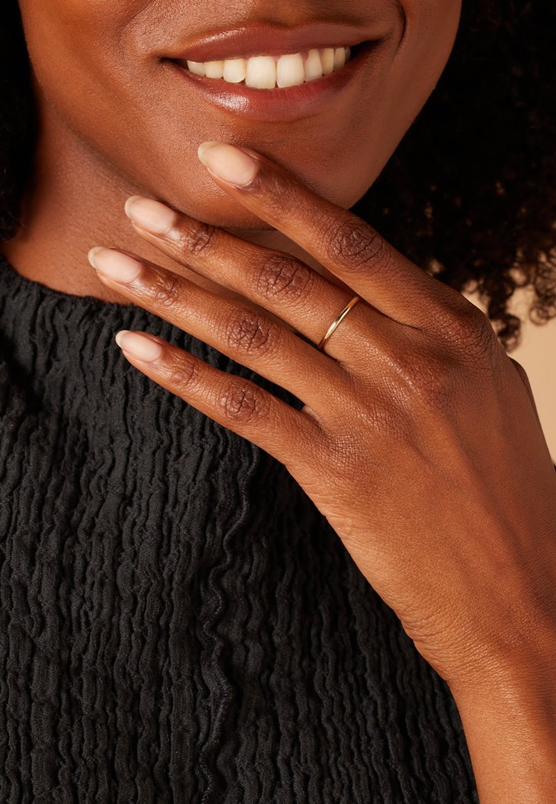 Gold ring on a finger resting on a textured black fabric. Smooth skin tone, natural nails with a light polish, and a warm smile visible.