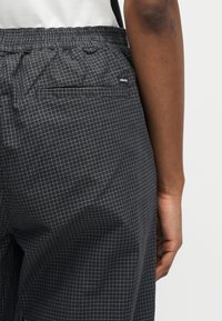 Άτομο που φοράει μαύρο και γκρι καρό παντελόνι με ελαστική μέση και πίσω τσέπη με την ετικέτα "obey", στέκεται μπροστά σε ένα απλό φόντο.