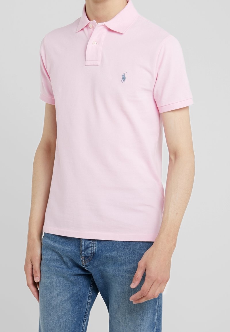 Homme portant un polo rose clair à manches courtes avec un logo brodé bleu associé à un jean bleu, sur fond uni.