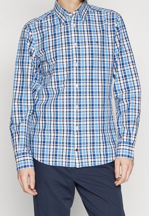 Homme portant une chemise à manches longues à carreaux bleus et blancs boutonnée et un pantalon bleu foncé, debout devant un fond uni.