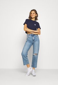 Mujer sonriendo con los brazos cruzados, vistiendo una camiseta corta azul marino, pantalones vaqueros de cintura alta desgastados y zapatillas altas blancas sobre un fondo blanco.