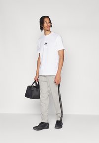 Witte katoenen t-shirt met zwart logo, grijze sweatpants met zwarte strepen, zwarte sport schoenen en een zwarte gymtas.