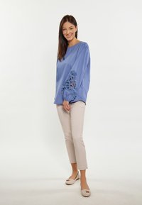 Blusa azzurra chiara con maniche a palloncino e dettagli in pizzo sui polsini, abbinata a pantaloni beige aderenti e ballerine beige con decorazioni.