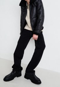 Musta nahkatakki, jossa on karvahihainen kaulus, vaaleankermanvärinen huppari alla, yhdistettynä mustiin suoralahkeisiin farkkuihin ja paksuihin mustiin kenkiin.