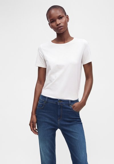 Femme portant un t-shirt blanc uni à manches courtes et un jean bleu, debout avec une main dans la poche devant un fond clair uni.