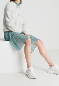 Bílý pletený svetr s žebrovanou texturou, spárovaný s teal vzorovanými šaty. Dokončeno s bílými sportovními teniskami.