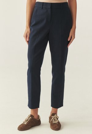 Vrouw die marineblauwe, op maat gemaakte enkelhoge broek en bruine suède veterschoenen draagt, staand tegen een neutrale achtergrond.