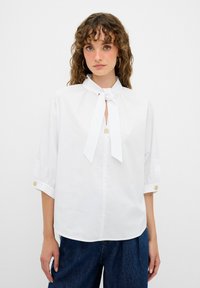 Hvid bluse med bånd omkring halsen, knapdetaljer på ærmerne og en løs pasform. Stoffet er glat med en let glans. Bæret med blå denim.