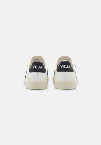 Hvite lær sneakers med en svart hælflapp, med "VEJA" merkevare og en kremfarget gummisåle. Minimalistisk design med rene linjer.