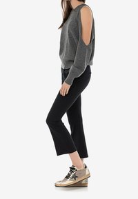 Kobieta stojąca bokiem, ubrana w szary sweter z odkrytymi ramionami, czarne spodenki 3/4 i beżowe trampki z czarnymi gwiazdkami.