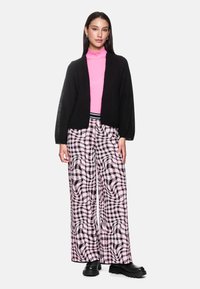 Frau in schwarzem Cardigan, rosa Rollkragenpullover, rosa und schwarzen weit geschnittenen gemusterten Hosen und schwarzen Plateauschuhen, die vor einem weißen Hintergrund steht.