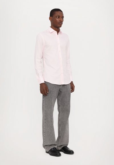 Camicia button-up rosa chiaro con colletto a punta, abbinata a pantaloni grigi strutturati e scarpe nere. Design semplice, vestibilità media.