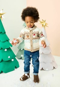 Kreminės ir smėlio spalvos megztinis su elnių ir sniego žmogelių raštais, derinamas su mėlynomis džinsomis ir rudomis botais. Fone matomi popieriniai kalėdiniai medžiai.