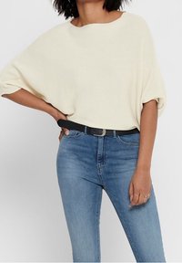 Jasno beżowy, dziergany, oversize sweter z szerokim dekoltem, zestawiony z wąskimi niebieskimi dżinsami i czarnym paskiem. Tekstura jest prążkowana.
