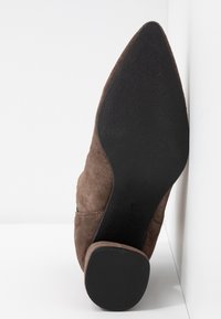 Chaussure à talon haut en daim marron avec un bout pointu et une semelle en caoutchouc noir texturé. Détails de couture visibles sur le côté.