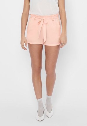 Femme portant un short taille haute rose clair avec une ceinture nouée, associée à des chaussettes en résille blanches et des escarpins blancs à bout pointu.