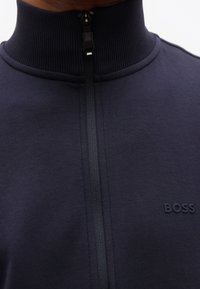 Κοντινό πλάνο ενός ατόμου που φοράει μπλε σκούρο πουλόβερ με φερμουάρ, ριμπ γιακά και το λογότυπο "BOSS" στο στήθος.