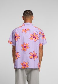 Korte ermers skjorte i lavendel med stort, oransje og rosa blomsterprint; stoffet virker lett og har en avslappet passform.