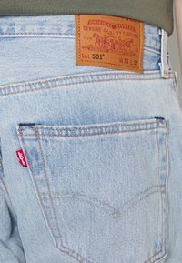 Light blue denim jeans featuring a brown leather patch with text, a red tab on the seam, and contrasting stitching on the back pocket.