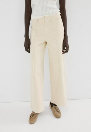 Persona con pantalones anchos color crema, top sin mangas blanco de canalé y zapatos beige de ante con cordones, de pie frente a un fondo blanco liso.