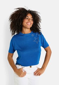 Suéter corto de punto azul con un brillo metálico y adornos de estrellas en el hombro izquierdo; combinado con pantalones blancos de cintura alta.