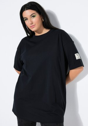 Vrouw met lang donker haar die een losse zwarte T-shirt draagt met een vierkante patch op de linkermouw, staand tegen een witte muur.