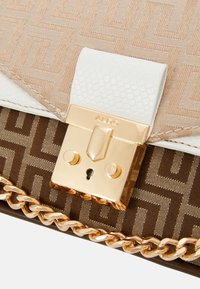 Gros plan d'un fermoir de sac à main avec une serrure dorée, cuir blanc texturé, tissu beige et marron à motifs, et une bandoulière chaîne dorée.