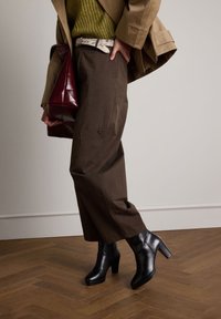 Braune Hosen mit weitem Bein, olivgrüner, gerippter Pullover, beiger Trenchcoat, schwarze Stiefeletten mit Absatz und eine dunkelrote Handtasche mit glänzendem Finish.