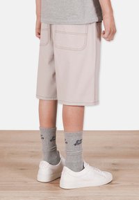 Ljusbeige shorts med två bakfickor och orange söm, parat med grå ribbade strumpor och vita sneakers på ett trägolv.
