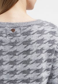 Primo piano della parte superiore della schiena di una persona che indossa un maglione a maglia di scozzese grigio e grigio chiaro, con una piccola etichetta in pelle marrone vicino al collo.