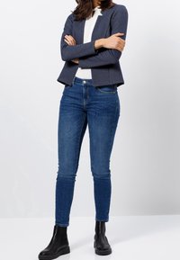 Femme portant un blazer bleu marine, un haut blanc, un jean skinny bleu et des bottines noires, debout les bras croisés devant un fond blanc.