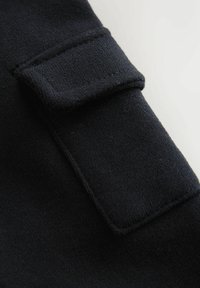 Schwarzer Stoff mit einer strukturierten Oberfläche, der ein rechteckiges Patch mit Nähten und einen gefalteten Überschlag für Details aufweist.